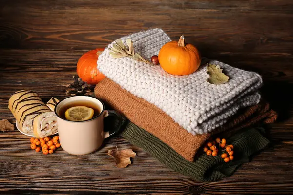 Sıcak kıyafetler, bir fincan çay, kek ve ahşap arka planda sonbahar dekoru.