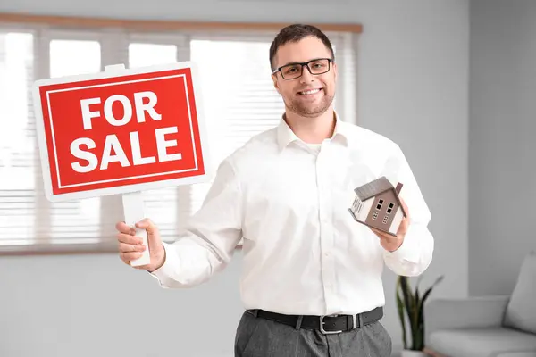 Male real estate agent with FOR SALE sign and house model in apartment