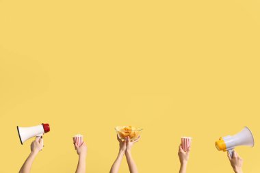 Patlamış mısır, patates cipsi ve sarı arka planda megafon bulunan kovaları tutan eller.