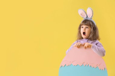 Şok olmuş küçük bir kız sarı arka planda dekoratif paskalya yumurtasıyla tavşan kulağı takıyor.