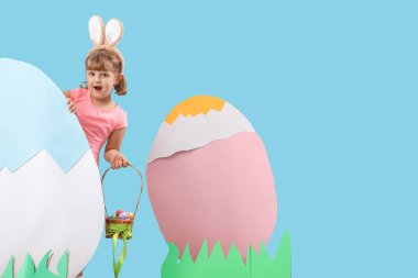 Şok olmuş küçük bir kız tavşan kulağı takıyor ve mavi arka planda Paskalya sepeti ve süslemeleri var.