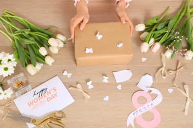 Hediye kutusu olan kadın elleri, güzel çiçekler, tebrik kartı ve tahta arka planda paketleme malzemeleri. Uluslararası Kadınlar Günü