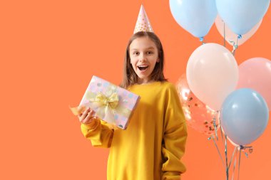 Parti şapkalı şirin bir kız. Doğum günü hediyesi ve turuncu arka planda balonlar.