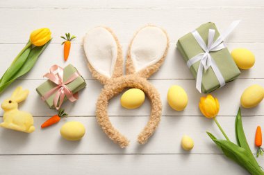 Paskalya yumurtalı tavşan kulakları, havuç, hediye kutuları ve beyaz ahşap arka planda lale çiçekleri.