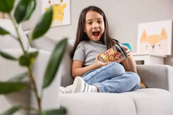 Cute little Asian girl with jar of cereal rings sitting on sofa at home