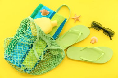 Hasır çantalı bir çift yeşil terlik ve sarı arka planda güneş kremi.