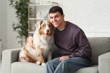 Genç adam evdeki kanepede Avustralyalı çoban köpeğine sarılıyor.
