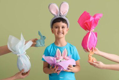 Tatlı küçük çocuk, tavşan kulağı takıyor, pasta yiyor ve kadın elleri yeşil arka planda Paskalya hediyelerini tutuyor.