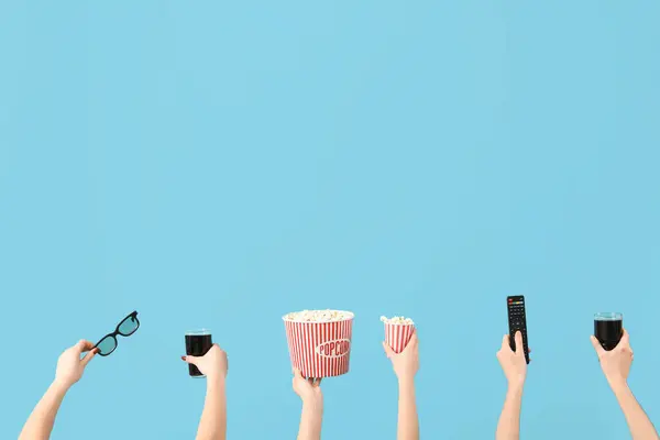 Hands holding buckets with popcorn, cola, 3D glasses and TV remote on blue background