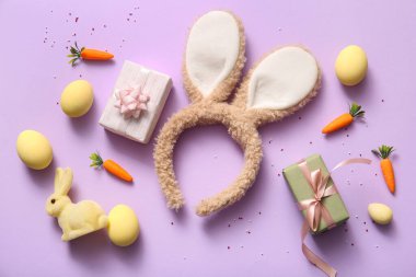 Paskalya yumurtalı tavşan kulakları, havuç ve leylak arkaplanda hediye kutuları.