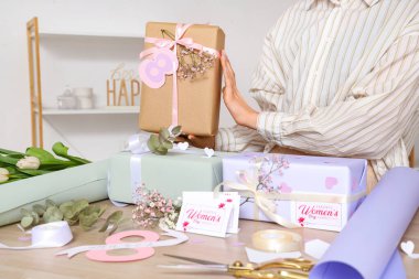 Güzel hediye kutuları, paketleme malzemeleri ve Uluslararası Kadınlar Günü için ışıklı duvarın yanındaki ahşap masada tebrik kartları olan bir kadın.