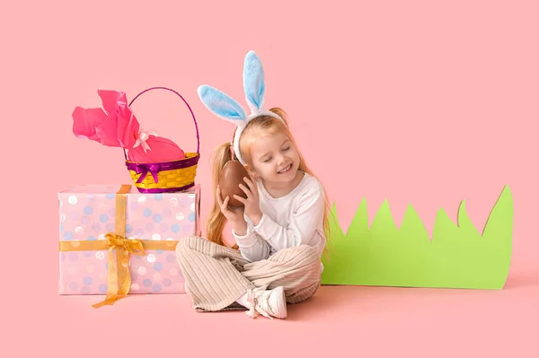在兔子耳朵里可爱的小女孩 戴着巧克力复活节彩蛋 礼物和粉红背景的纸草 — 图库照片