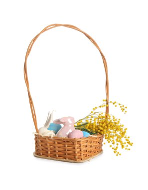 Beyaz arka planda boyanmış Paskalya yumurtaları, seramik tavşanlar ve çiçeklerle dolu bir sepet.