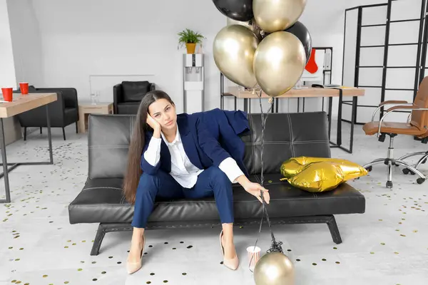 Hangover businesswoman with balloons sitting on couch after New Year party in office