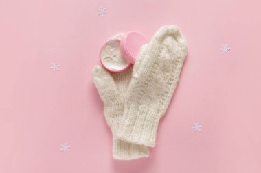 Kar taneli bir kavanoz krema ve pembe arka planda sıcak kış eldivenleri.