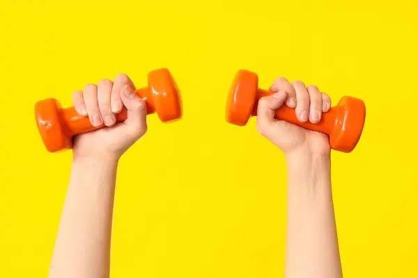 Female hands with orange dumbbells on yellow background, closeup