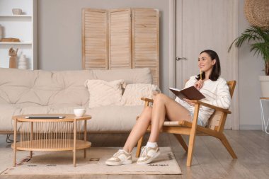 Evdeki tahta koltukta oturan defterli mutlu genç kadın.