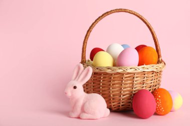 Renkli Paskalya yumurtaları ve pembe arka planda tavşanlı hasır sepet.