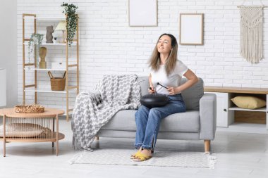 Güzel olgun bir kadın sopayla glukofon çalıp evdeki kanepede oturuyor.