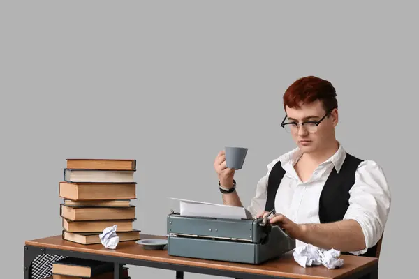 Young man with vintage typewriter and cup of coffee at table on grey background