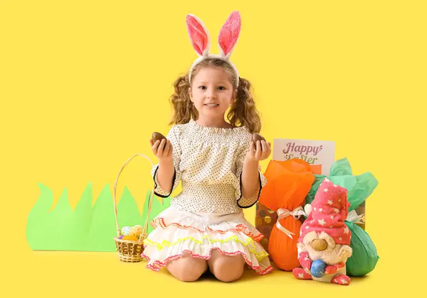 可爱的小女孩 长着小兔子耳朵 戴着巧克力复活节彩蛋 坐在黄色的背景上 — 图库照片