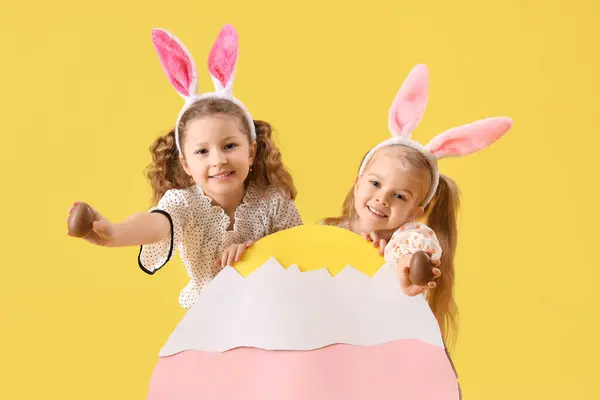 用巧克力复活节彩蛋和黄色底色的纸装饰兔子耳朵里可爱的小女孩 — 图库照片