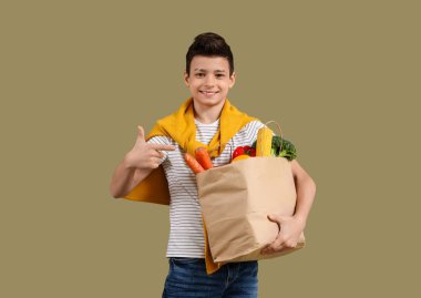Yeşil arka planda farklı sebzelerle dolu kağıt torbayı işaret eden sevimli küçük çocuk.