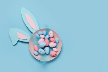 Mavi arka planda Paskalya yumurtası, tüy ve tavşan kulaklı tabak.