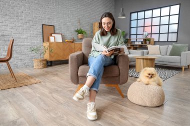 Dergisi olan mutlu genç bir kadın ve Pomeranya tükürüğü evde koltukta oturuyor.