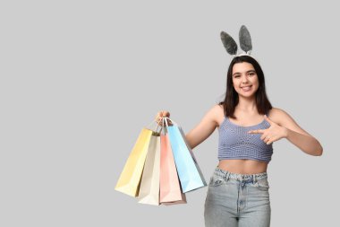 Tavşan kulaklı güzel, genç ve mutlu bir kadın gri arka plandaki alışveriş çantalarını işaret ediyor. Paskalya kutlaması