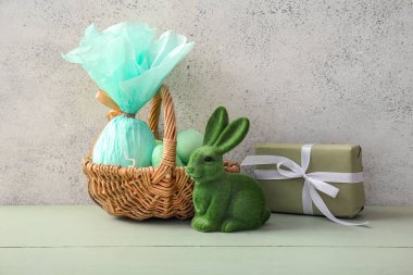 Boyanmış Paskalya yumurtaları, hediye çantası ve yeşil ahşap masada oyuncak tavşancık olan hasır sepet.