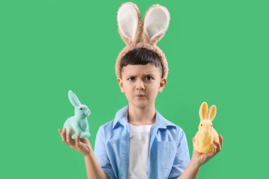 Kafası karışmış küçük çocuk tavşan kulağı takıyor ve yeşil arka planda Paskalya tavşanları var.