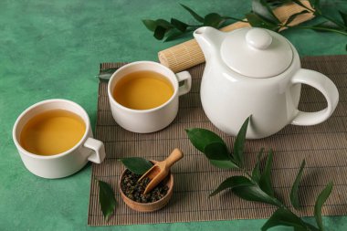 Çaydanlıklı bambu hasırı, bir fincan çay ve yeşil grunge arka planda yapraklar.