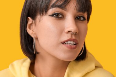 Sarı arka planda burun piercingli şık bir genç kadın.