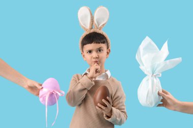 Çikolata yumurtalı tavşan kulağı takan düşünceli küçük çocuk ve mavi arka planda Paskalya hediyelerini tutan kadın elleri.