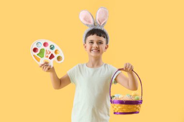 Paskalya sepetiyle tavşan kulağı takan ve sarı arka plan boyalı şirin bir çocuk.