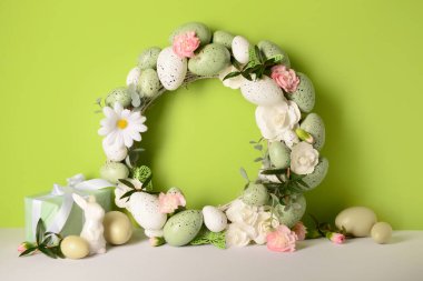 Küçük tavşan, şirin bir hediye, yeşil arka planda çiçekler ve yumurtalarla süslenmiş Paskalya çelengi.