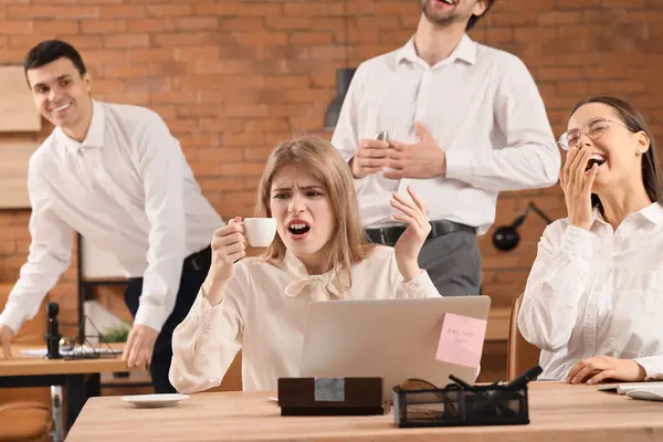 Angry young woman with salty coffee and her laughing colleagues in office. April Fools\' Day celebration