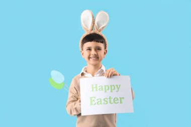 Paskalya hediyesi kartıyla tavşan kulağı takan şirin bir çocuk ve mavi arka planda kağıt yumurta.