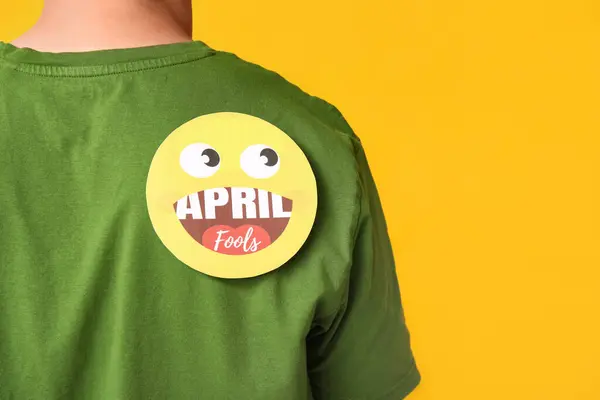 Handsome young man with paper smile attached to his back on yellow background. April fool\'s day celebration
