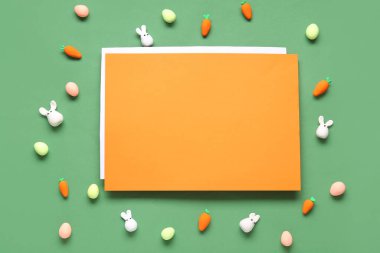 Paskalya tavşanları, boyalı yumurta, havuç ve yeşil arka planda turuncu boş kart. Üst görünüm