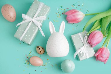 Paskalya hediyeleri, yumurtalar, tavşan figürü ve mavi arka planda laleler.