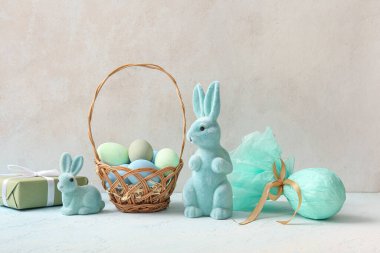 Hasır sepet, boyalı Paskalya yumurtaları, hediye paketi ve arka planda oyuncak tavşanlar.