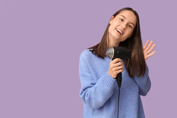 Beautiful young woman with hair dryer on lilac background