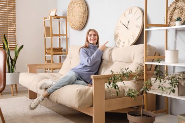 Oturma odasındaki kanepenin üzerindeki büyük saati gösteren genç kadın.