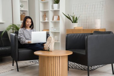 Genç Asyalı kadın evdeki siyah koltukta dizüstü bilgisayar kullanıyor.