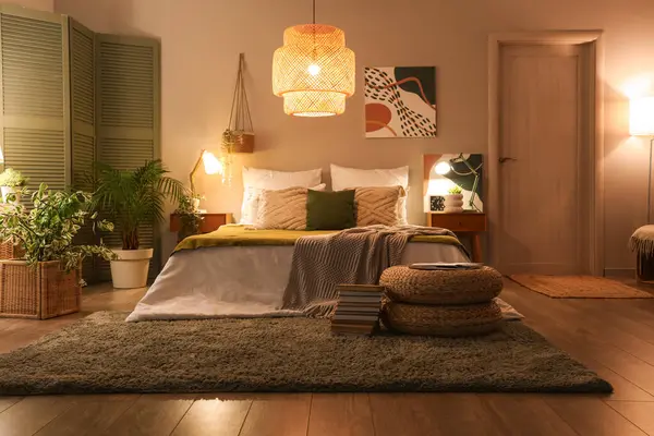 Interior of stylish bedroom with blankets on bed, wicker poufs and glowing lamps at night