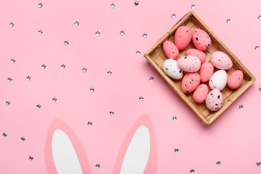 Paskalya yumurtaları ve pembe arka planda kağıt tavşan kulakları olan bir kutu.