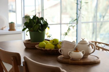 Mutfağın içinde yemek masası, koltuklar, ev bitkileri, bulaşık takımı ve elmalar.