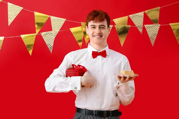 Young man with piece of birthday cake and gift on red background